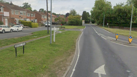 A road junction on a quiet road with houses to the left and large grass verges