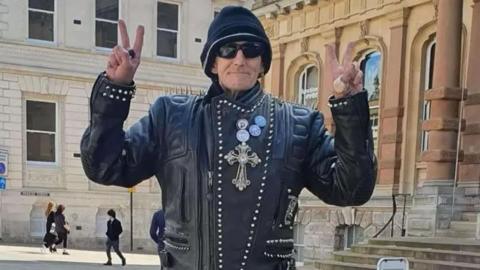 William McNicholl is pictured outside the Town Hall in Ipswich. He is wearing a black leather jacket and making 'peace' signs with both of his hands. He's also wearing sunglasses and a hat