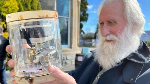 Loch Ness expert Adrian Shine is an older man with white hair and a long white beard. He is wearing a blue jacket and is holding the camera. The camera is inside a tube-like glass container.