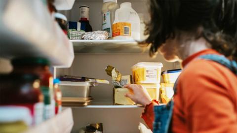 Someone with short dark hair and an orange t-shirt opens a fridge and reaches inside. The fridge is quite full and well stocked with dairy products like milk and butter.
