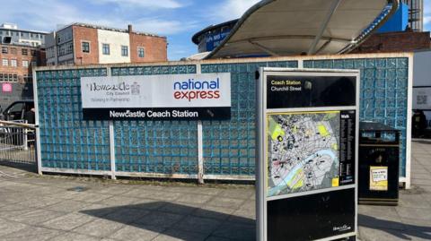 Newcastle Coach Station. A wall made from glass squares stands in front of the coach stop. On it is a sign stating that Newcastle City Council and National Express are working in partnership with each other. A map of Newcastle city centre stands in the foreground.