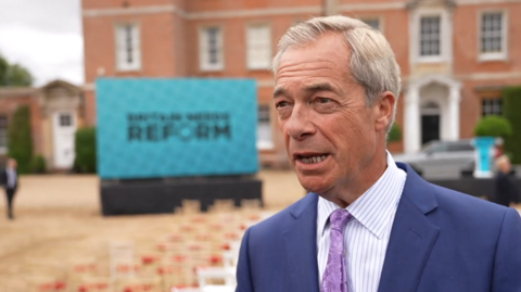 Nigel Farage looks towards a reporter off camera, with his mouth open as he talks, while wearing a blue suit with a striped shirt and purple tie, with chairs outside a stately home blurred in the background
