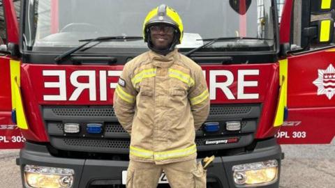 Zekel Johnson in his uniform and a helmet, with his hands behind his back, standing in front of a fire engine.