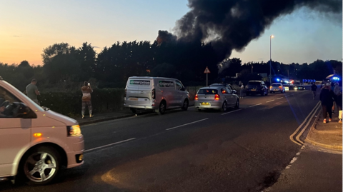 Thick black smoke rises above a road lined with parked vehicles