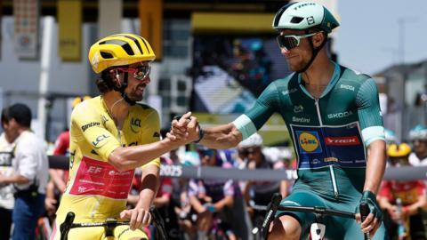 Two cyclists taking part in the Tour de France are sitting on their bikes and shaking hands. EasyPost's Ben Healy is wearing the yellow jersey and smiling while he shakes hands with Lidl - Trek's Jonathan Milan wearing the green jersey before stage 11 in 2025.