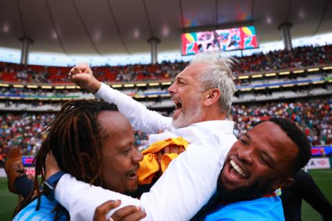 DR Congo coach Sebastien Desabre is seen in a long-sleeved smart white shirt being lifted up by two players, with one grabbing him from behind and the other from in front. Desabre is punching the air and screaming with joy as he celebrates his side's World Cup qualification. In the background can be seen the high tiers of a stand with red seats