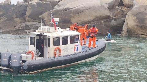 Taiwanese coast guard personnel dressed in orange uniforms on board a coast guard ship apprehend a suspect dressed in a blue top and grey trousers, who appears to be kneeling on a small floating platform. The vessels are by large rocks.