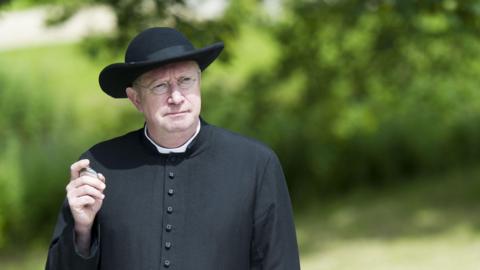 Mark Williams as Father Brown. He is dressed in black clerical robes and a wide-brim hat. He is holding a silver pocked watch and is also wearing thin, silver glasses. 