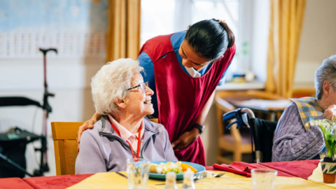 A woman with dark hair wearing a red tabard over a blue top is leaning over an elderly woman who is eating a plate of food at a table.