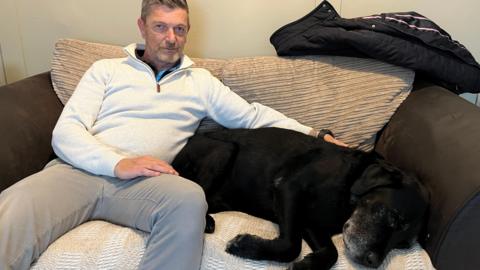 A man wearing a cream jumper and grey trousers is sat on a sofa with a black Labrador, greying around the muzzle, 