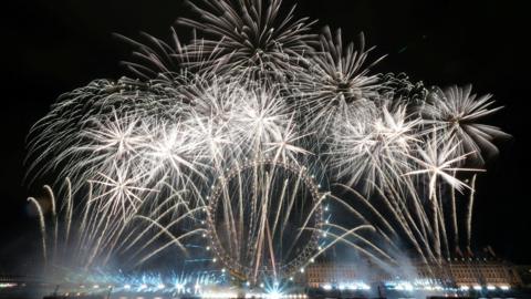 The night sky is filled with exploding fireworks all around the London Eye. More fireworks are being fired from a barge in the Thames in front of the Eye. 