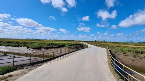 View of the smooth, resurfaced road, which looks like a bridge over a river with flat green fields in the distance and a blue sky