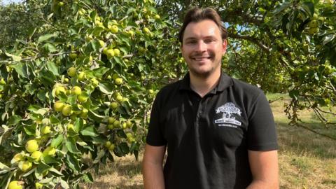 George Scott is standing in front of an apple tree at an orchard. He is wearing a black T-Shirt and is smiling.
