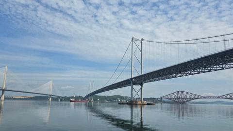 The Queensferry Crossing, Forth Road Bridge and Forth Rail Bridge spanning across the water. The reflections of the bridges are in the water and there are clouds in the sky.