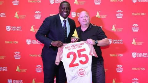 Emile Heskey stands with Darren Richards in front of a red board. The pair are holding a white t-shirt with the words New Volunteer of The Year and the number 25 written in red. Mr Richards is also holding a trophy in his right hand.