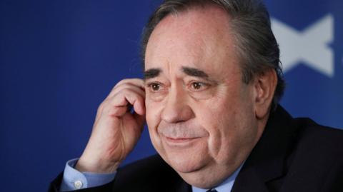 Alex Salmond in close up against a saltire background. He is wearing a dark jacket, mid-blue shirt, and is smiling.