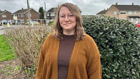 Merryn Tenalach stands in a street in front of bushes. She is wearing glasses, a brown top and brown woollen cardigan.
