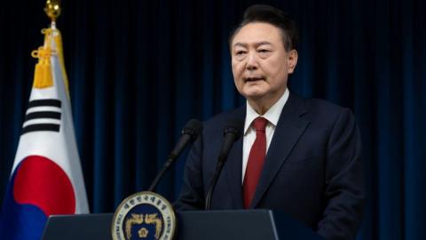 Yoon Suk Yeol wearing a navy suit and red tie speaking at a podium. A large South Korean flag is beside him.