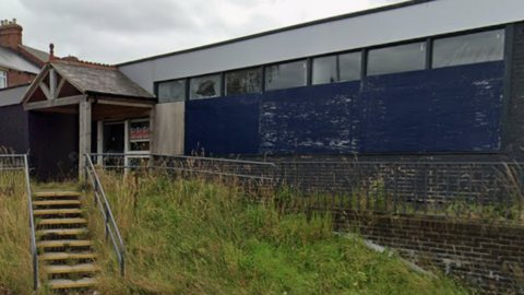 The former Stanley library is an empty one-storey building made of brown bricks and surrounded by overgrown grass. 