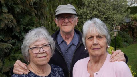 Two women with short hair, they are standing in front of a man, who is wearing a hat. They appear to be standing in a garden. Close up photo. One of the women and the man are wearing glasses.