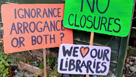 Three banners reading 'ignorance, arrogance or both?', 'we love our libraries' and 'no closures' can be seen in the photo.