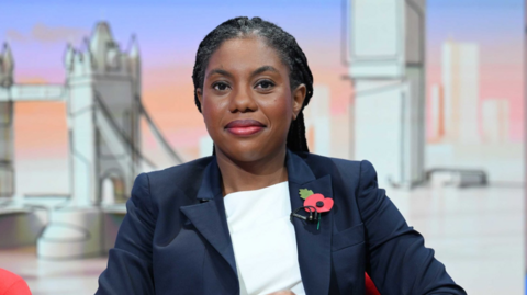 Kemi Badenoch dressed in a navy blue blazer and a white top. She is wearing a poppy on her blazer and is looking directly at the camera. She is in a TV studio and behind her you can see sketch style drawings of London landmarks