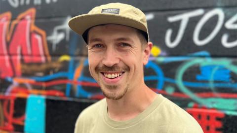 Head and shoulders shot of Shawn Sharpe standing in front of a graffiti wall. He is wearing a beige cap and t-shirt, has a short dark goatee and is smiling