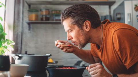 A man leans near a pot on the stove with a wooden spoon to his mouth, tasting the food he is cooking.