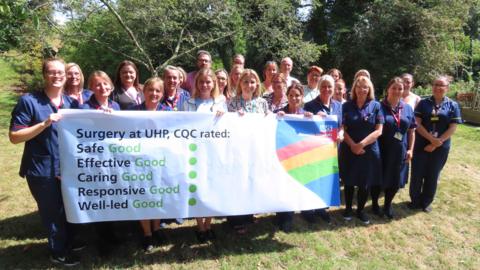 Staff from Derriford Hospital stand on a lawn in front of trees and hold up a banner saying CQC has rated surgery services at the site as good. The word good is written five times in green along with five green dots. Many of the staff are wearing scrubs.