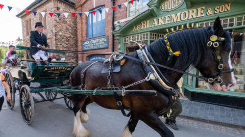 A horse drawn cart driven by a man in a black suit and top hat with a large brick building in the background with a painted green shop front and a sign reading AF Blakemore and son
