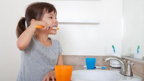 A girl brushes her teeth in front of a sink. There are blue and orange plastic beakers on the surface and there is another toothbrush in a holder by the taps. She is looking into the mirror as she cleans her teeth.