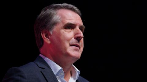 Steve Rotheram looking out to the distance. He is wearing a dark blazer jacket with an open top button shirt