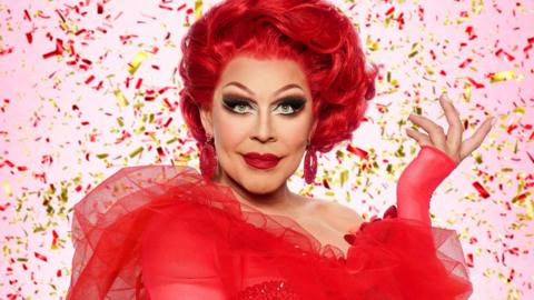La Voix, with a thick red mane of hair and wearing a red dress, stands against a pink background surrounded by gold and red confetti
