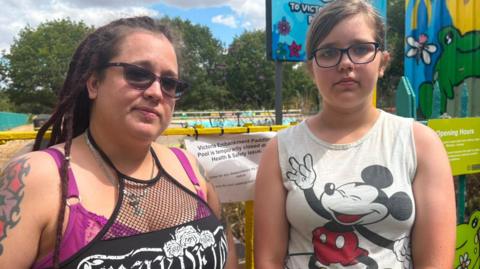 A mother and daughter, both wearing glasses stand in front of a locked gate to the Victoria Embankment Paddling Pool. There is a colourful temporary storage building behind the girl and the drained paddling pool beyond.