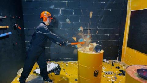 A woman smashing a table full of crockery and electrical items placed on a yellow container in a small room . She is wearing blue overalls and an orange hat with visor.