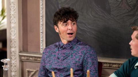 TV still of James during an episode of Traitors. He is sitting at a banquet table looking towards a fellow contestant. His mouth and eyes are wide open as if he is in shock. He is wearing a purple and black marble shirt and he has short black curly hair.