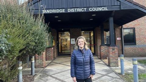 Tandridge District Council leader Catherine Sayer wearing a blue puffer jacket. She is stood in front of a red brick building with a black wooden awning. The awning has a white sign reading "Tandridge District Council" on it.