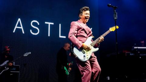 Rick Astley on stage at Bestival with a sign saying "Astley" behind him, he holds a guitar and wears a pink suit