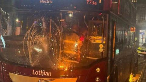 The windscreen of the Lothian bus is smashed. The bus is maroon and gold.