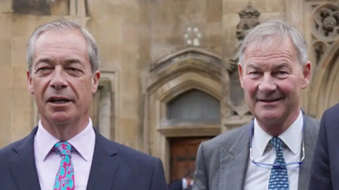 On the left is Nigel Farage who has grey hair, and is wearing a navy suit, with a pink shirt, and pink and blue tie. On the right is Rupert Lowe, who has grey hair, a grey suit jacket, white shirt, and blue tie. He has glasses around his neck. 