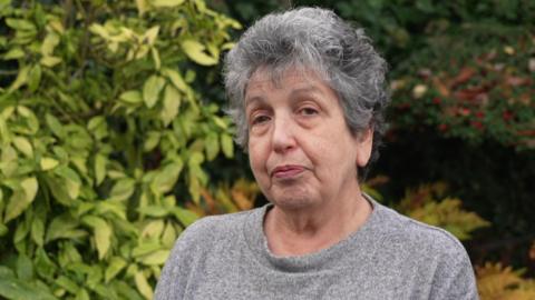Ruth Jacobs, a woman with grey hair, wearing a grey jumper, is standing outdoors. There is a backdrop of trees.
