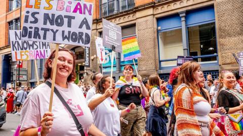 A Pride parade scene in Manchester shows people marching along the street holding signs and rainbow flags. One prominent sign reads “Lesbian is not a dirty word”. Colourful outfits and banners fill the lively crowd against a backdrop of brick buildings.