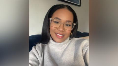 A woman with dark hair wearing glasses and a grey sweater sits smiling at the camera. Her hair is past her shoulders and she sits on a blue couch in front of a white wall.