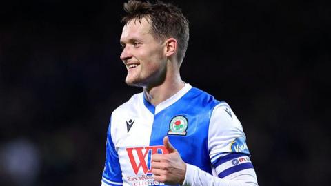 Blackburn Rovers defender Sean McLoughlin smiling with left thumb up looking to his right in home kit