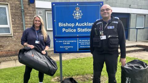 A man and a woman, smiling, stand outside a police station next to a blue sign which reads 'Bishop Auckland Police Station'.
To the left, a woman with long blonde hair, wearing a dark blue hoodie, holds a big black duffel bag with the words 'Sleep Pod'; to the right, stands a man wearing glasses and a police uniform - there is a Sleep Pod, also in a duffel bag, on the ground beside him.