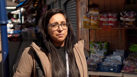 A woman wearing glasses and in a beige coat stands infront of a market stall selling crisps