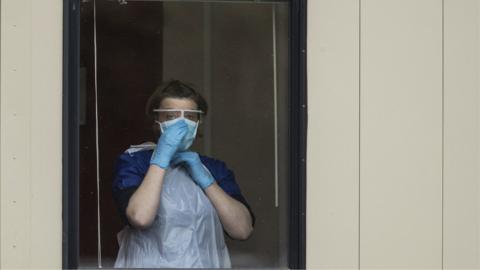 An NHS worker in PPE standing in front of a window