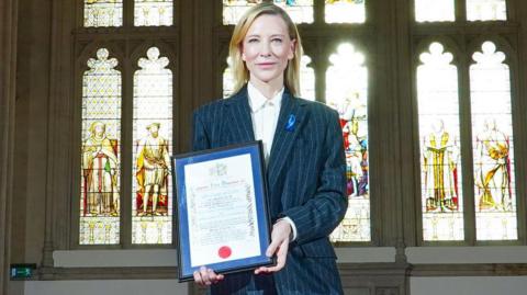 Cate Blanchett, a woman with long, blonde hair, wears a grey suit and hold up a framed certificate. She stands in front of a row of stained glass windows with light pouring in.