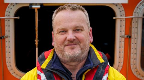 Steve Bennetts smiling at the camera. He is wearing a red and yellow jacket. He is standing in front of a lifeboat.