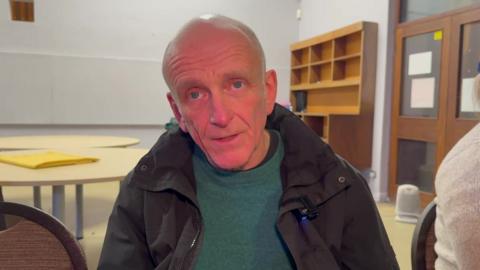 Gary Spencer, who is wearing a black coat and green jumper, is sitting on a chair during a choir singing session. 